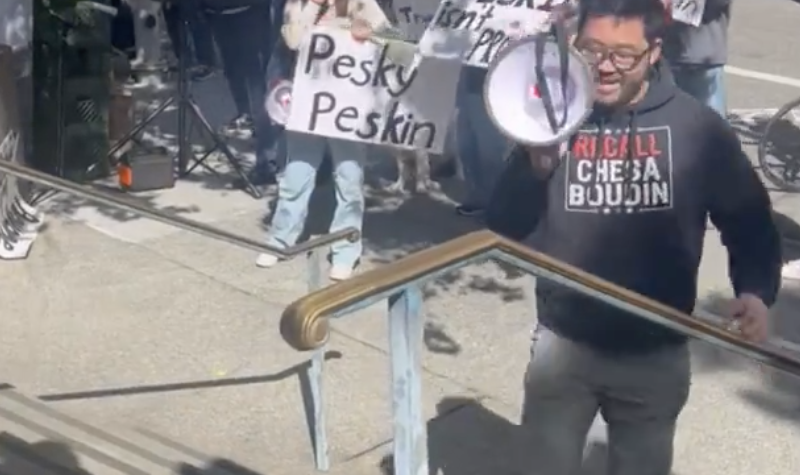 A person holds a megaphone while others stand with signs in a protest on a city sidewalk.